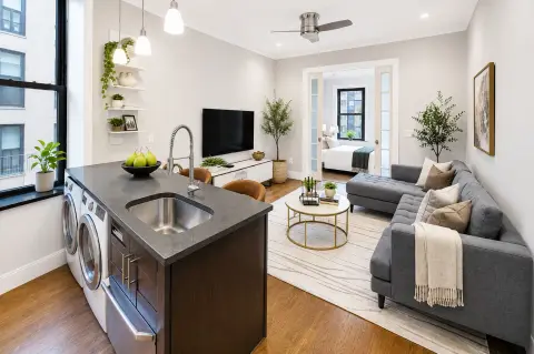 Open-plan kitchen and living room with dark island, stainless steel appliances, gray sofa, TV, plants, and hardwood floors.