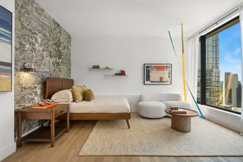 Bedroom with wooden bed frame, nightstand, beige rug, wall art, floating shelves, and floor-to-ceiling windows with city views.