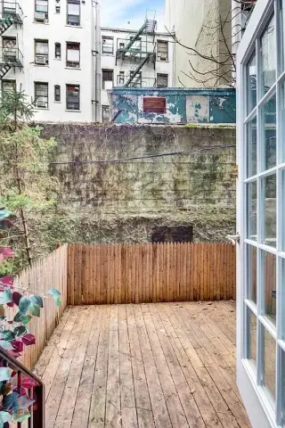 Outdoor deck with wooden flooring, brown fence, potted plants, and view of surrounding buildings.