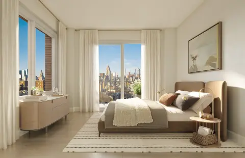 Bedroom with large windows overlooking city skyline, beige bed with pillows, dresser, and area rug.