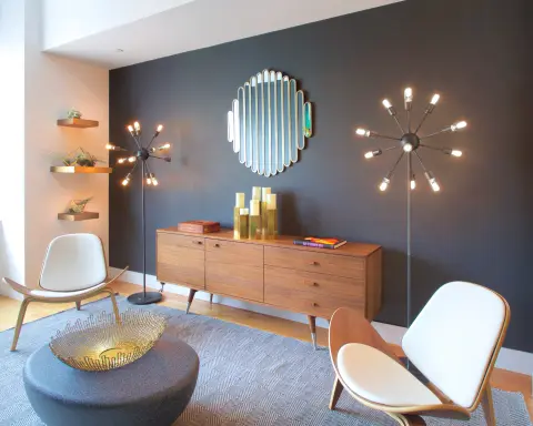 Living room with dark blue accent wall, wooden dresser, round ottoman, two modern chairs, and decorative wall sconces and mirror.