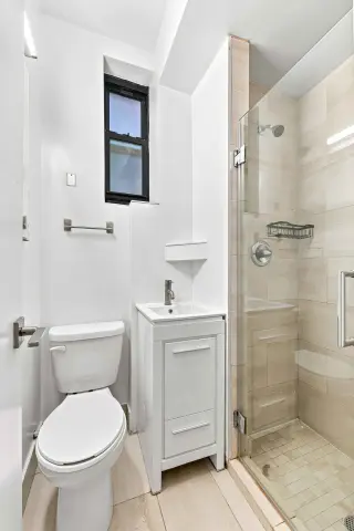 Bathroom with white toilet, small vanity sink, window, and glass-enclosed shower with beige tile walls.