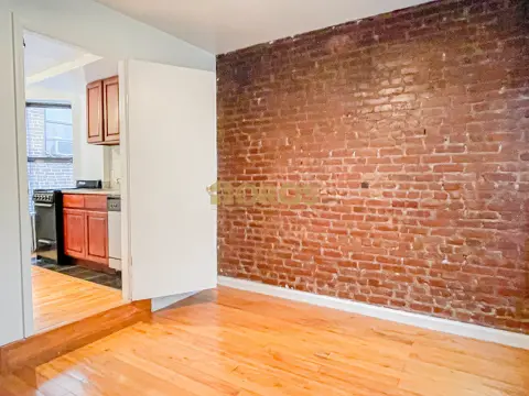 Living room with hardwood floors, exposed brick wall, and open doorway to kitchen with orange cabinetry.