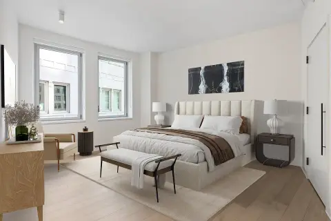 Bedroom with cream upholstered bed, two large windows, wooden dresser, nightstands, and light wood flooring.