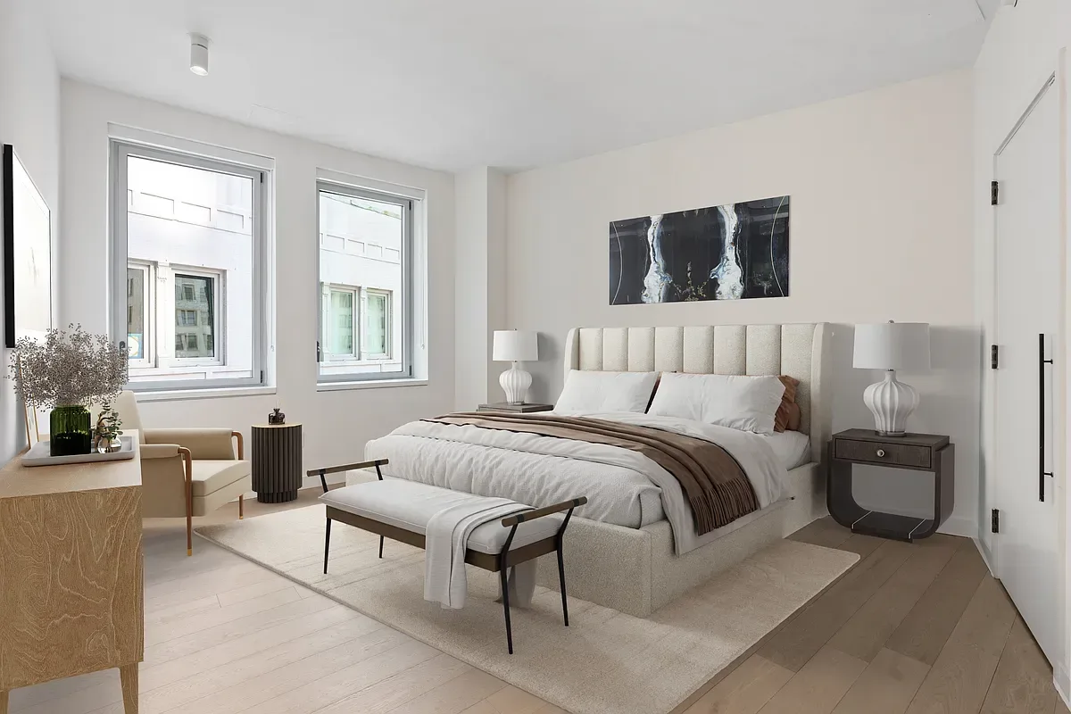 Bedroom with cream upholstered bed, two large windows, wooden dresser, nightstands, and light wood flooring.