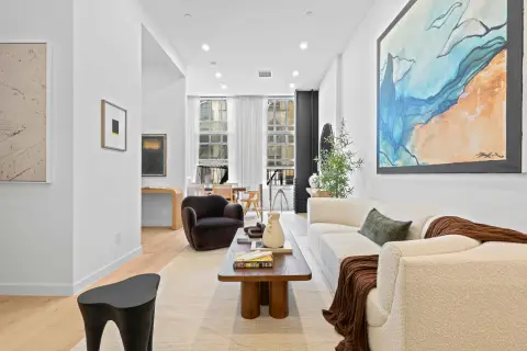 Open-plan living room with cream sofa, wooden coffee table, hardwood floors, large windows, and abstract artwork on walls.