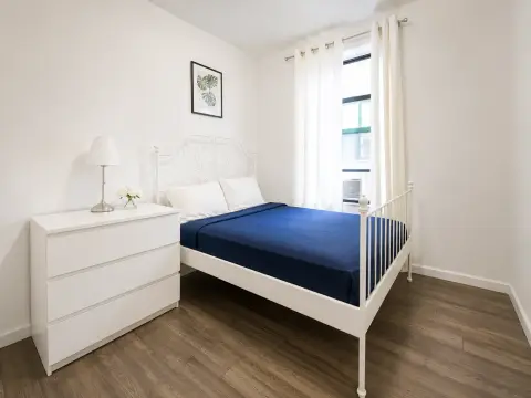 Bedroom with white bed frame, blue mattress, white dresser, bedside lamp, and window with blue shutters.