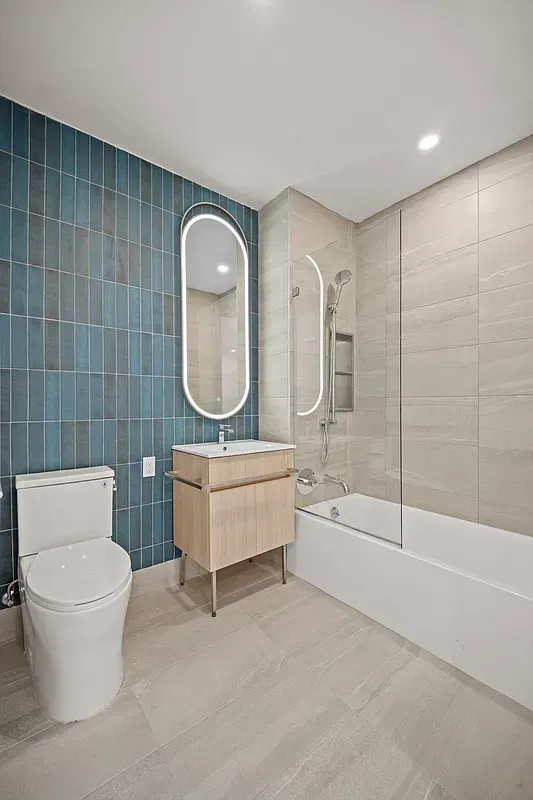 Modern bathroom with teal tile accent wall, white vanity, oval mirror, toilet, and bathtub with glass enclosure.
