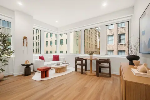 Bright living room with hardwood floors, large windows overlooking city buildings, white seating, wooden furniture and potted plants.