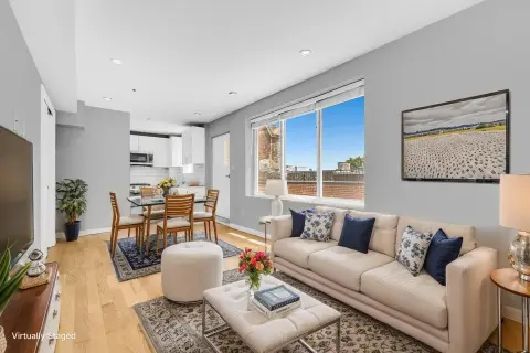 Open-plan living and dining area with hardwood floors, beige sofa, dining table, large windows with city views, and framed artwork on walls.