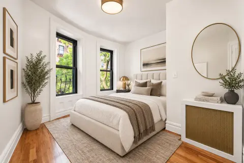 Bedroom with cream upholstered bed, hardwood floors, two black-framed windows, round mirror, wooden dresser and potted plants.