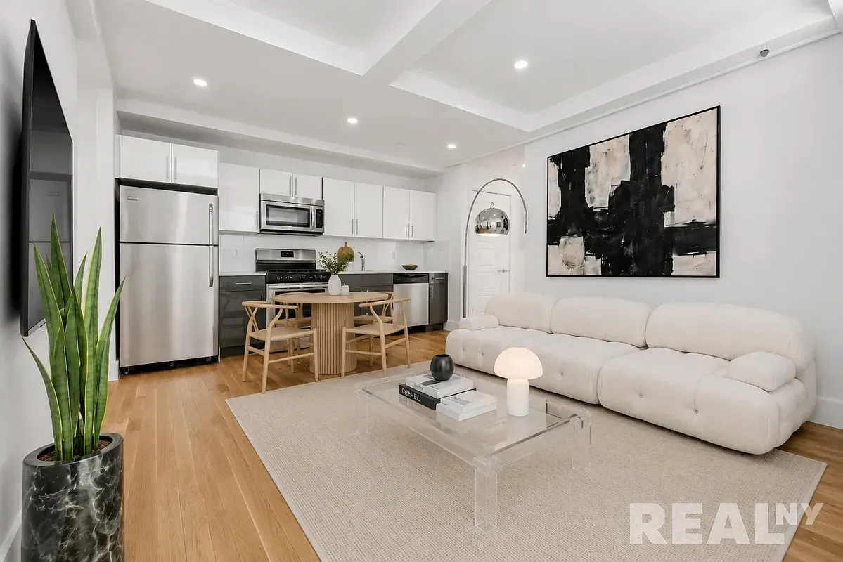 Open-concept living room and kitchen with white cabinetry, stainless steel appliances, wooden dining table, cream sofa, and hardwood floors.