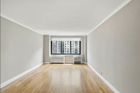 Empty room with white walls, wooden floor, and large window overlooking city buildings.
