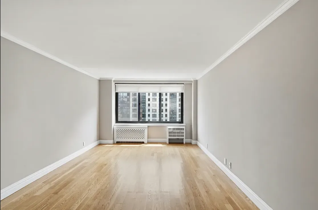 Empty room with white walls, wooden floor, and large window overlooking city buildings.